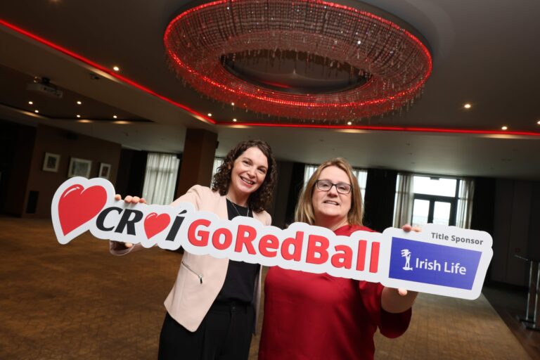 Two smiling women holding a 'Croí Go Red Ball' sign, standing under a large chandelier in a well-lit event space.