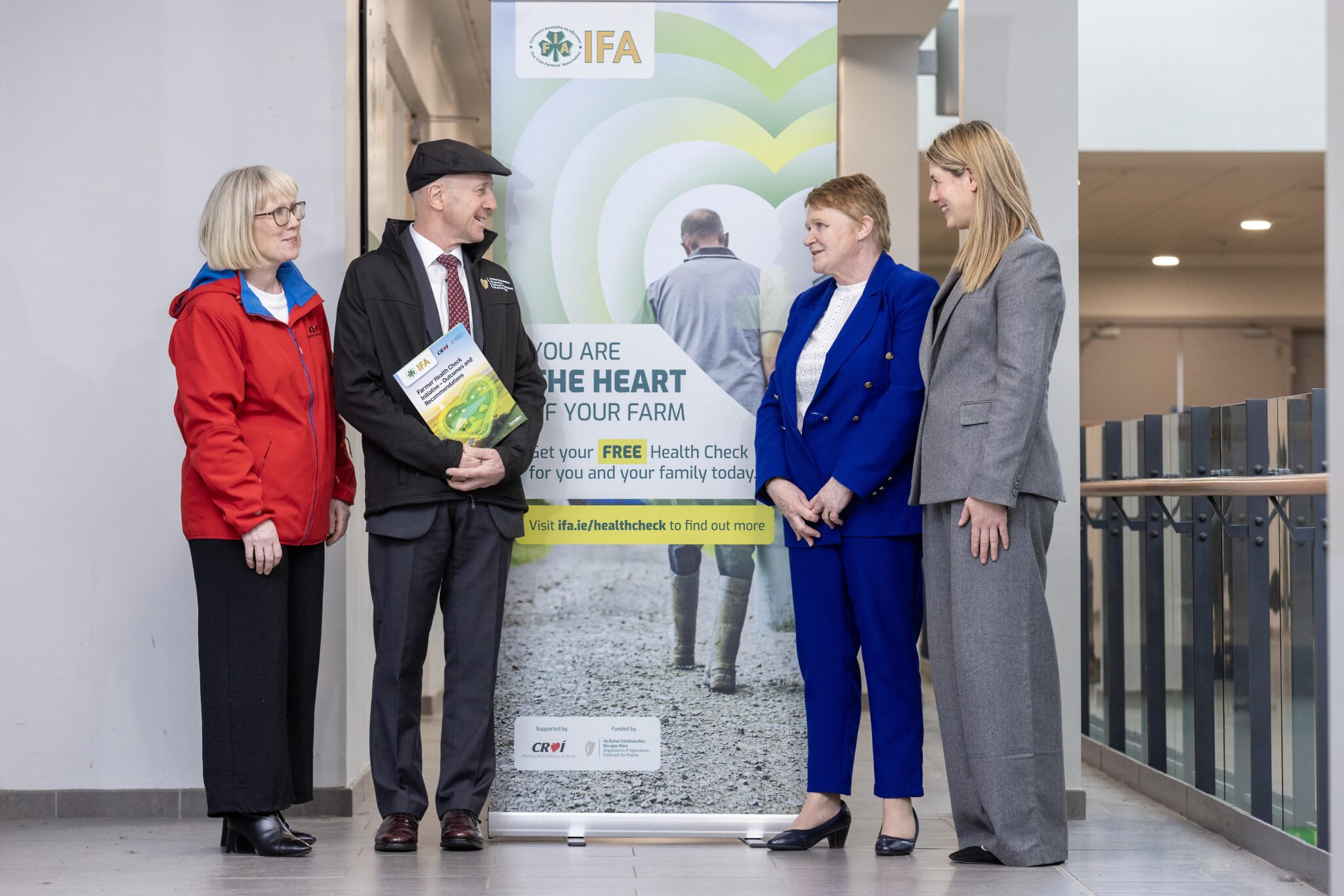 Four people standing in front of a banner promoting free health checks for farmers, including one man holding a booklet talking to three women/