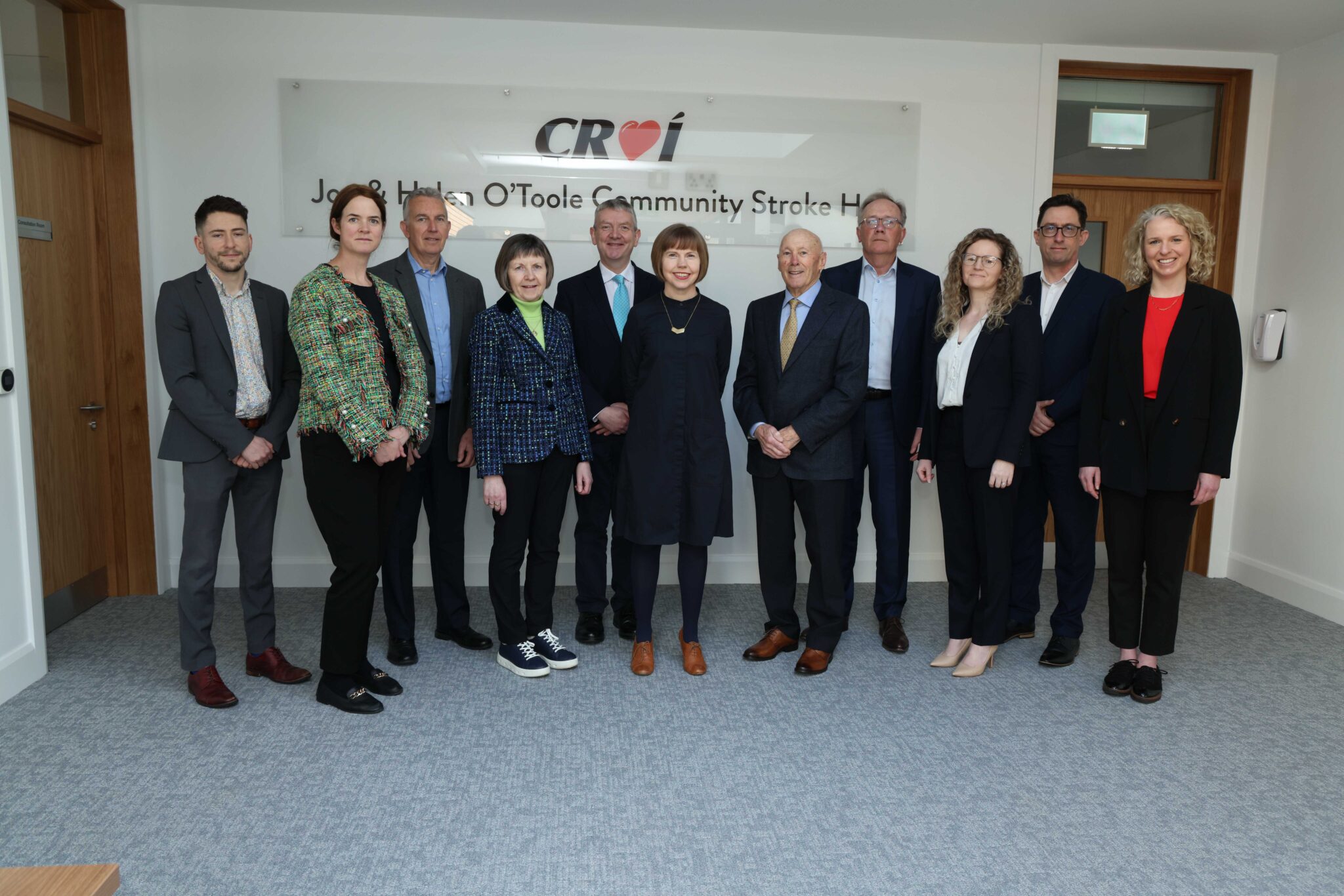 A group of ten professionally dressed individuals standing in a well-lit office space posing for a photo in front of a sign that reads 'Croí Joe & Helen O'Toole Community Stroke Hub'.