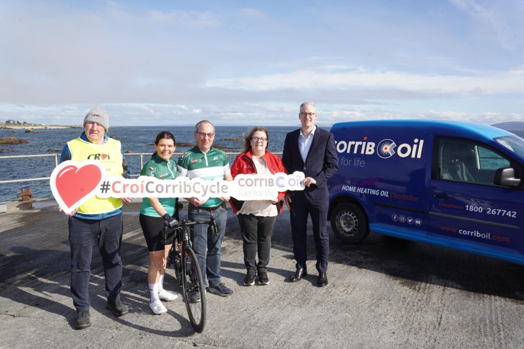A group of five people posing near the seaside, with a Corrib Oil branded van in the background, holding a sign that reads '#CroíCorribCycle'. One person is wearing cycling gear and holding a road bike.