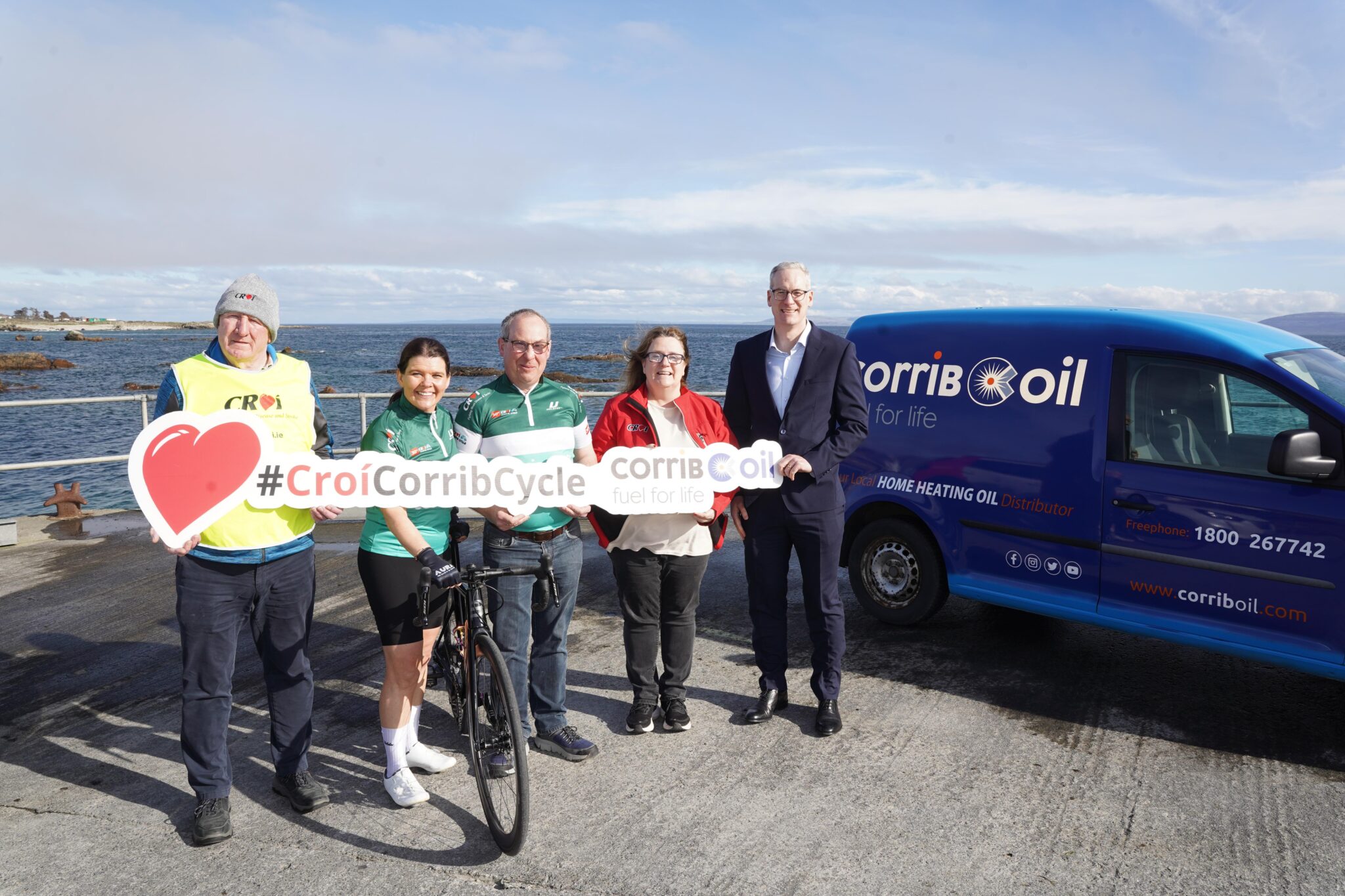 A group of five people posing near the seaside, with a Corrib Oil branded van in the background, holding a sign that reads '#CroíCorribCycle'. One person is wearing cycling gear and holding a road bike.