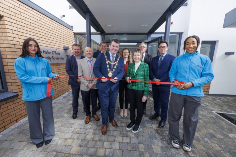A group of people stand in front of the Joe & Helen O'Toole Community Stroke Hub at the Croí Heart & Stroke Centre in Galway for a ribbon-cutting ceremony. A man wearing a ceremonial chain holds scissors alongside a woman in a green plaid blazer, preparing to cut the red ribbon. Two individuals in blue Croí-branded jackets hold the ribbon at each end, while several formally dressed attendees stand behind them, smiling.