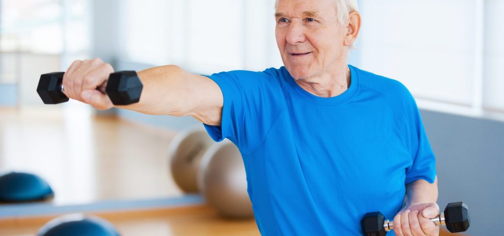 Confident senior man exercising with dumbbells and smiling while standing in health club